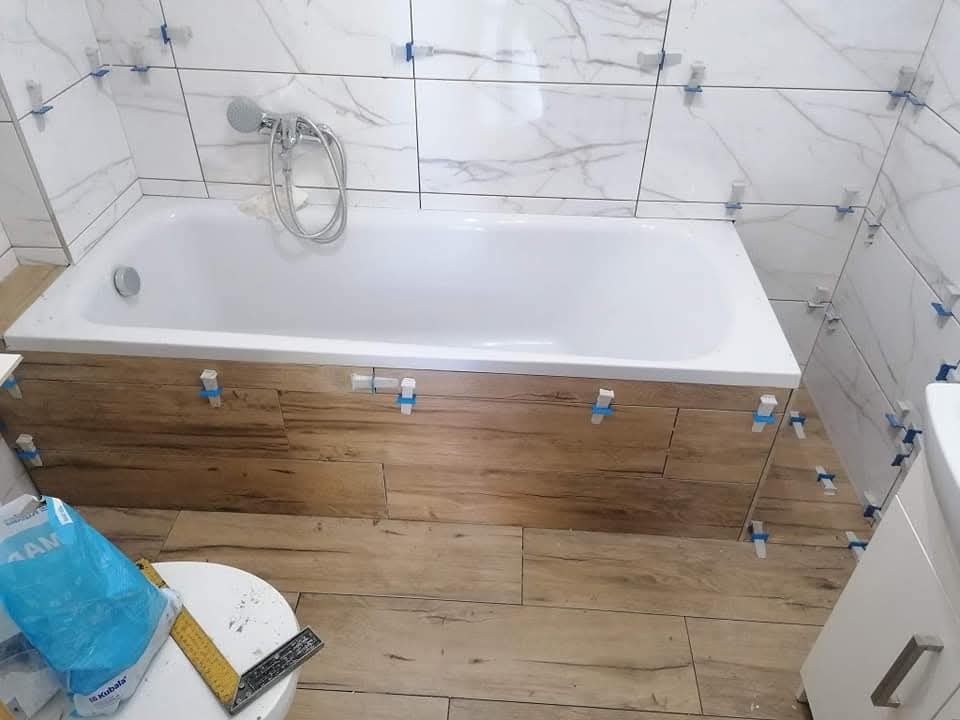 Montaż płytek imitujących drewno na obudowie wanny i podłodze w łazience. Widoczne krzyżyki dystansowe i białe płytki marmurowe na ścianach. Etap prac wykończeniowych.