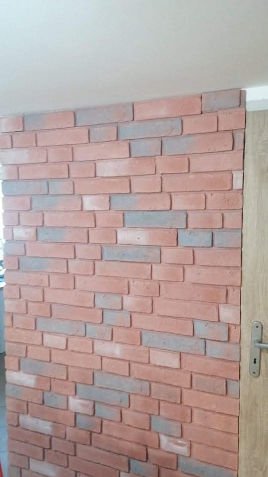 Ściana wykończona dekoracyjną cegłą w odcieniach czerwieni i szarości, przylegająca do drewnianej ościeżnicy drzwiowej z widoczną klamką. Nowoczesny design wnętrza.