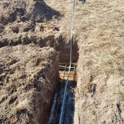 Wykop pod fundament z widocznym zbrojeniem stalowym, pryzma ziemi w tle, widoczny fragment pojazdu.