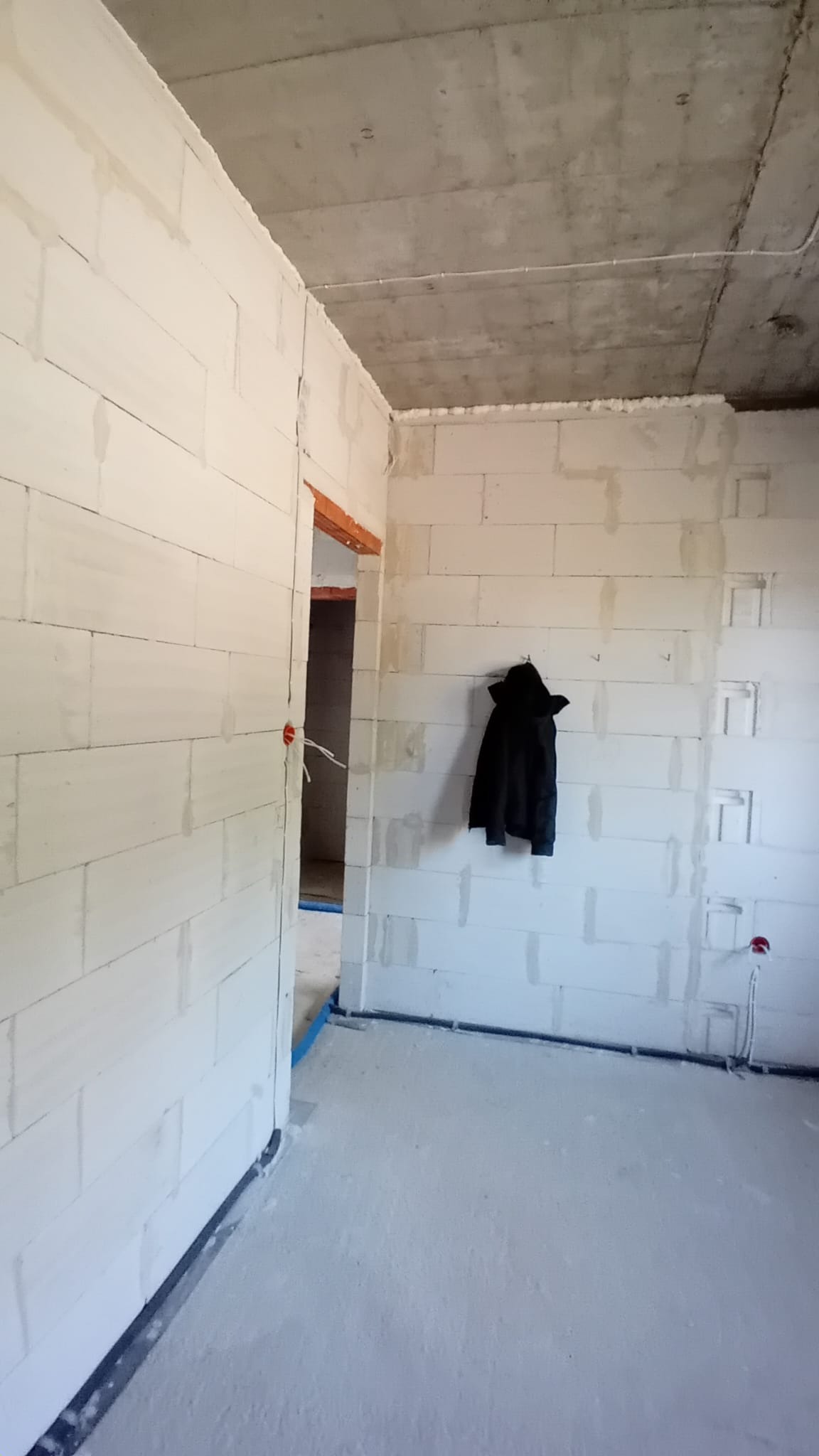 Surowy stan wnętrza w trakcie prac wykończeniowych: widoczne ściany z bloczków, betonowy sufit z przewodami, wystające kable ze ściany, czarna kurtka wisząca na ścianie.