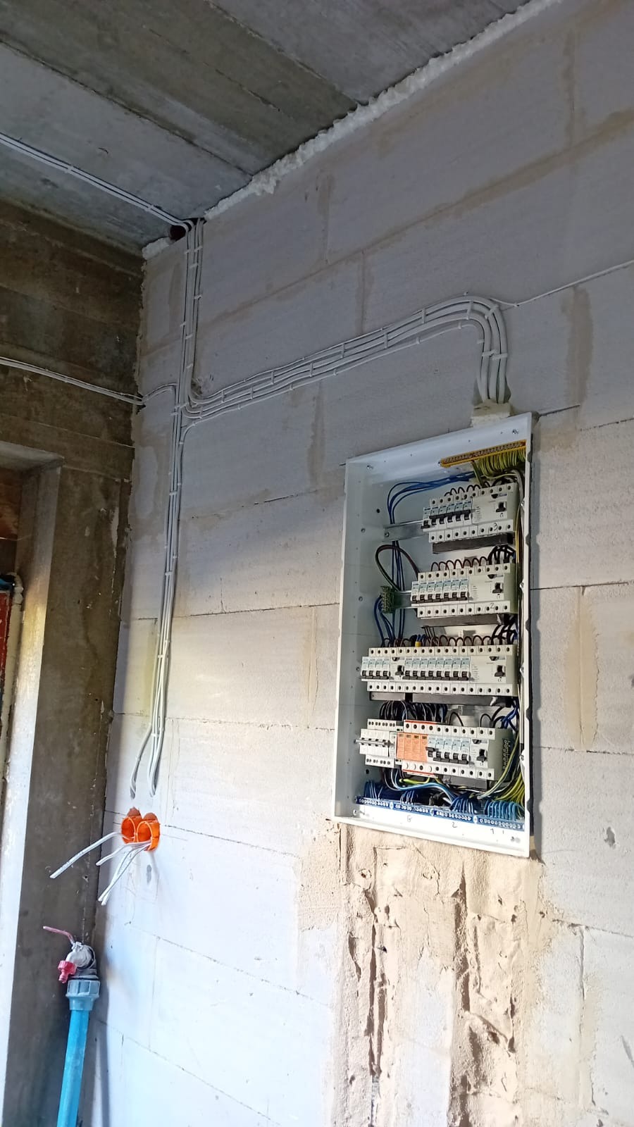 Rozdzielnica elektryczna z zabezpieczeniami zamontowana na ścianie z betonu komórkowego, widoczne kable w białych peszlach, puszka instalacyjna z wystającymi przewodami. W tle surowy betonowy strop.