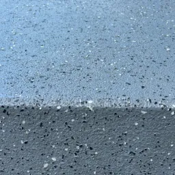 Szara powierzchnia z drobnymi, jasnymi i ciemnymi elementami, przypominająca lastryko, z widoczną krawędzią.