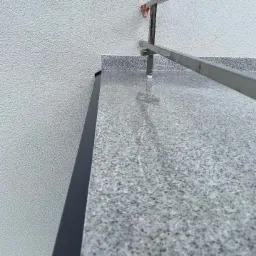 Szara, drobnoziarnista powierzchnia balkonu wykończona czarną listwą, z fragmentem metalowej balustrady i białą, chropowatą ścianą w tle.