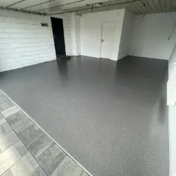 Szara, gładka posadzka w garażu, z widocznym fragmentem kostki brukowej na zewnątrz, białe ściany i sufit z widocznymi elementami konstrukcyjnymi.