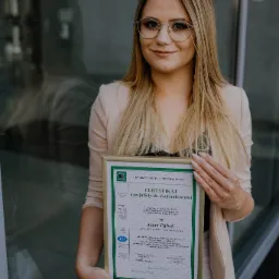 Uśmiechnięta blondynka w okularach prezentuje certyfikat specjalisty ds. rachunkowości na tle szklanej fasady budynku.