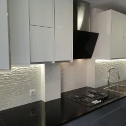 Nowoczesna kuchnia z białymi szafkami, czarnym blatem, płytą grzewczą i trójwymiarową białą mozaiką na ścianie.