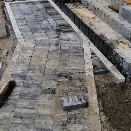 Układanie chodnika z szarej kostki brukowej, z widocznym poziomowaniem i przygotowaniem podłoża, w tle schody z kostki i narzędzia budowlane.