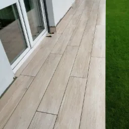 Wyłożony płytkami taras z widokiem na trawnik, płytki imitujące drewno, widoczne dystanse do płytek przy oknie.