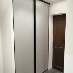 Wnękowa szafa przesuwna z matowymi, szarymi drzwiami i czarnymi obramowaniami w białym korytarzu z szarą podłogą, oświetlona liniowym LED-em na suficie, obok ciemnobrązowe drzwi.