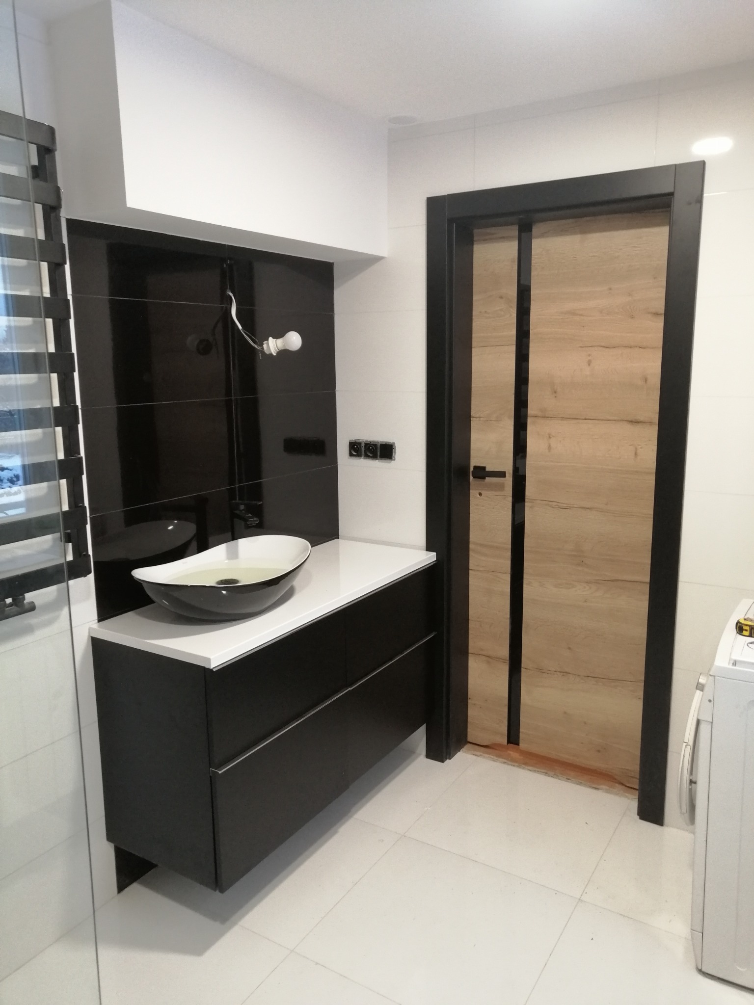 Nowoczesna łazienka z czarną armaturą, umywalką w kształcie misy i drzwiami z drewnianym fornirem w czarnej ramie. Widoczna biała glazura i pralka.