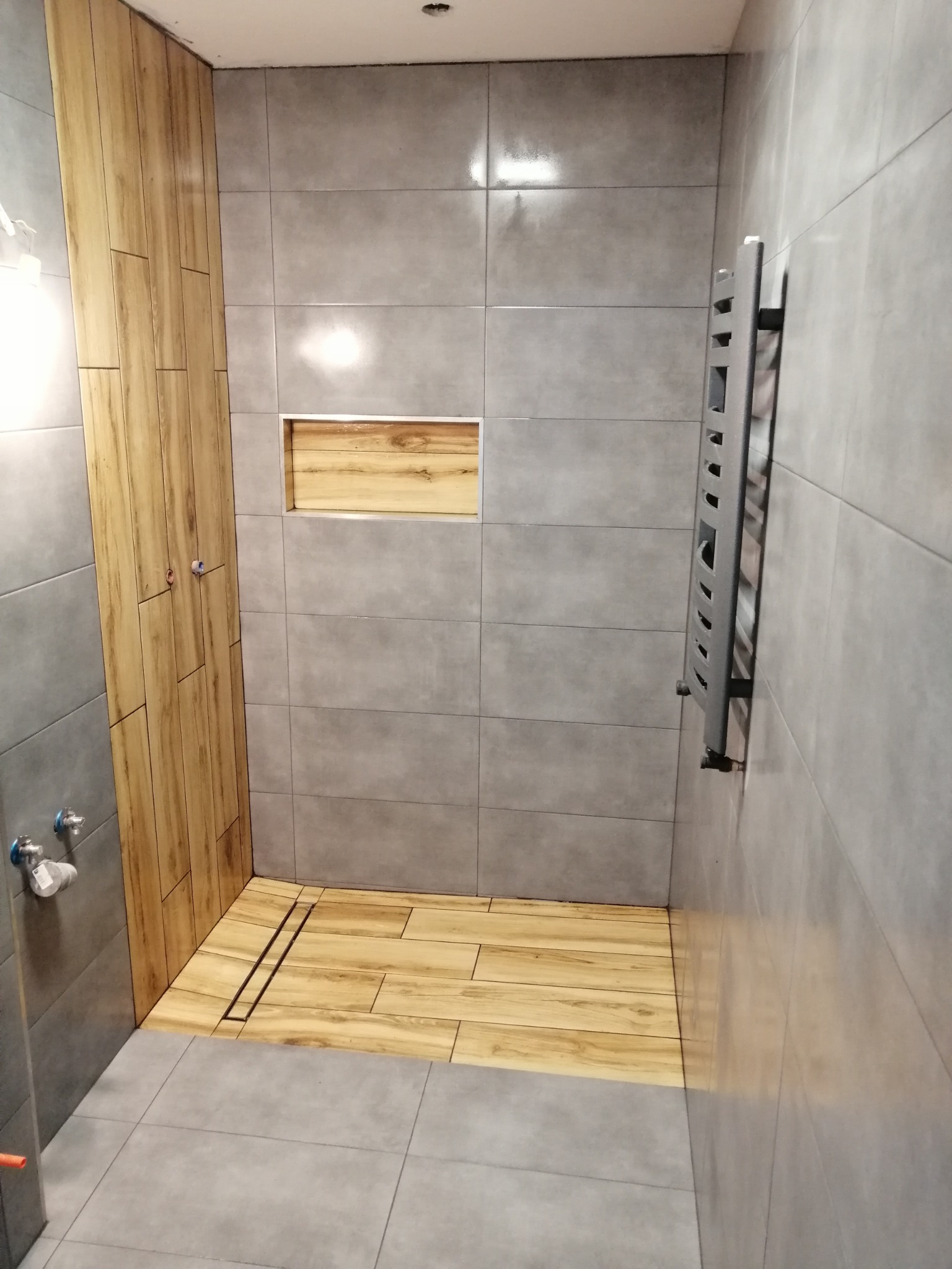 Nowoczesna łazienka z szarymi płytkami i akcentami drewna: drewnopodobna podłoga i ściana kabiny prysznicowej, wnęka ścienna z drewnianym wykończeniem, grzejnik drabinkowy.