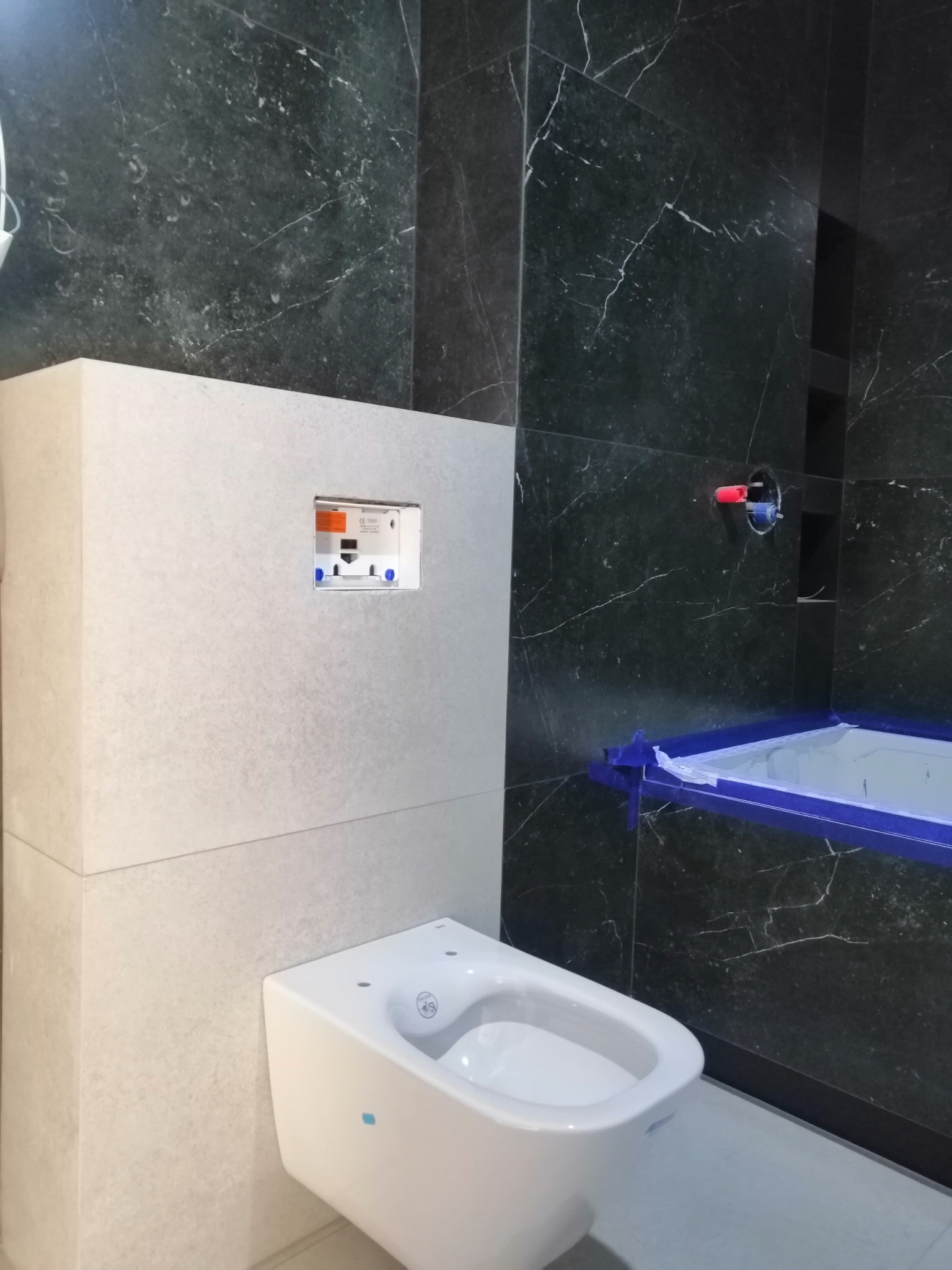 Wnętrze łazienki w trakcie montażu: wisząca toaleta, obudowa stelaża z otworem na przycisk spłukujący, czarne płytki imitujące marmur, zabezpieczona folią wanna.