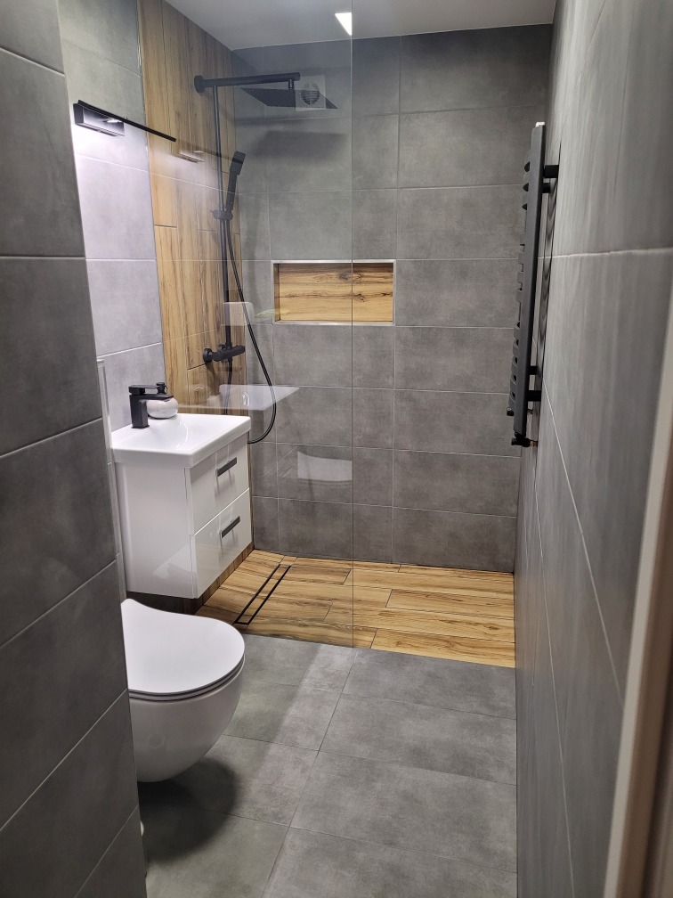 Nowoczesna łazienka z szarymi płytkami, kabiną prysznicową typu walk-in z czarną armaturą, drewnopodobną wnęką i podłogą prysznicową, białą szafką z umywalką i wiszącą toaletą.