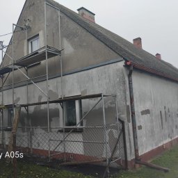 MATEUSZ GIŻA STACHSTON ELEWACJE - Stary dom w trakcie remontu elewacji. Widoczne rusztowania, częściowo otynkowana ściana i fragment dachu. Ujęcie z dołu, pochmurna pogoda.