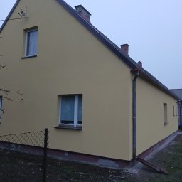 MATEUSZ GIŻA STACHSTON ELEWACJE - Żółta elewacja domu jednorodzinnego z białymi oknami i szarymi rynnami, widok z boku, ogrodzenie z siatki w tle, pochmurne niebo.