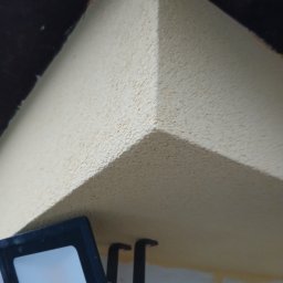 MATEUSZ GIŻA STACHSTON ELEWACJE - Wykończony narożnik sufitu z fakturą baranka, oświetlony lampą roboczą. Widoczne niedoskonałości malarskie przy ścianie.