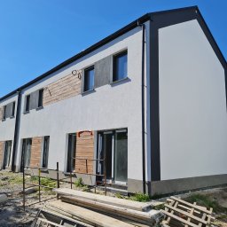 MATEUSZ GIŻA STACHSTON ELEWACJE - Nowoczesny budynek po ociepleniu, z fragmentami drewnianej elewacji i rusztowaniami w tle, w słoneczny dzień. Widoczne palety i drabina.