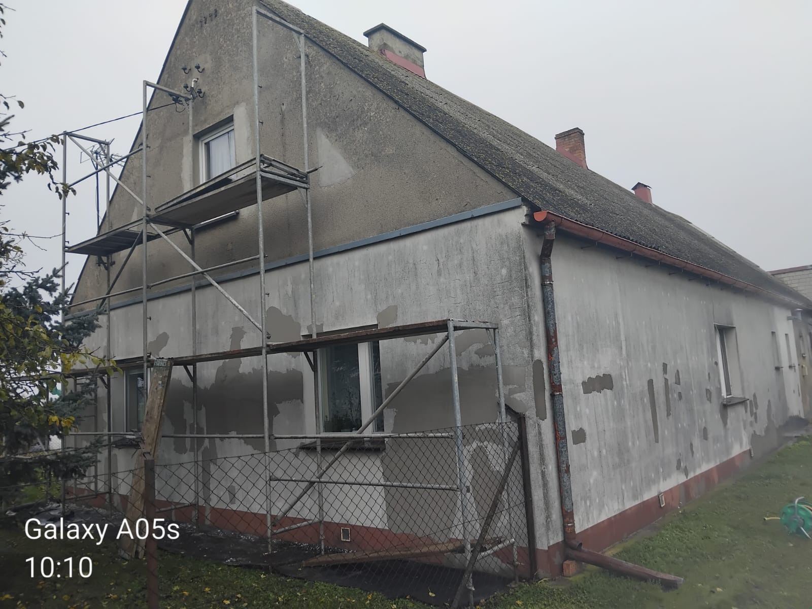 Stary dom w trakcie remontu elewacji. Widoczne rusztowania, częściowo otynkowana ściana i fragment dachu. Ujęcie z dołu, pochmurna pogoda.