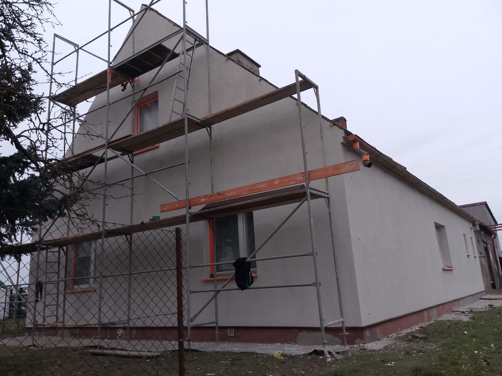 Ocieplony dom jednorodzinny w trakcie prac wykończeniowych, z widocznymi rusztowaniami i zabezpieczonymi oknami. Świeżo nałożony tynk w odcieniach szarości.