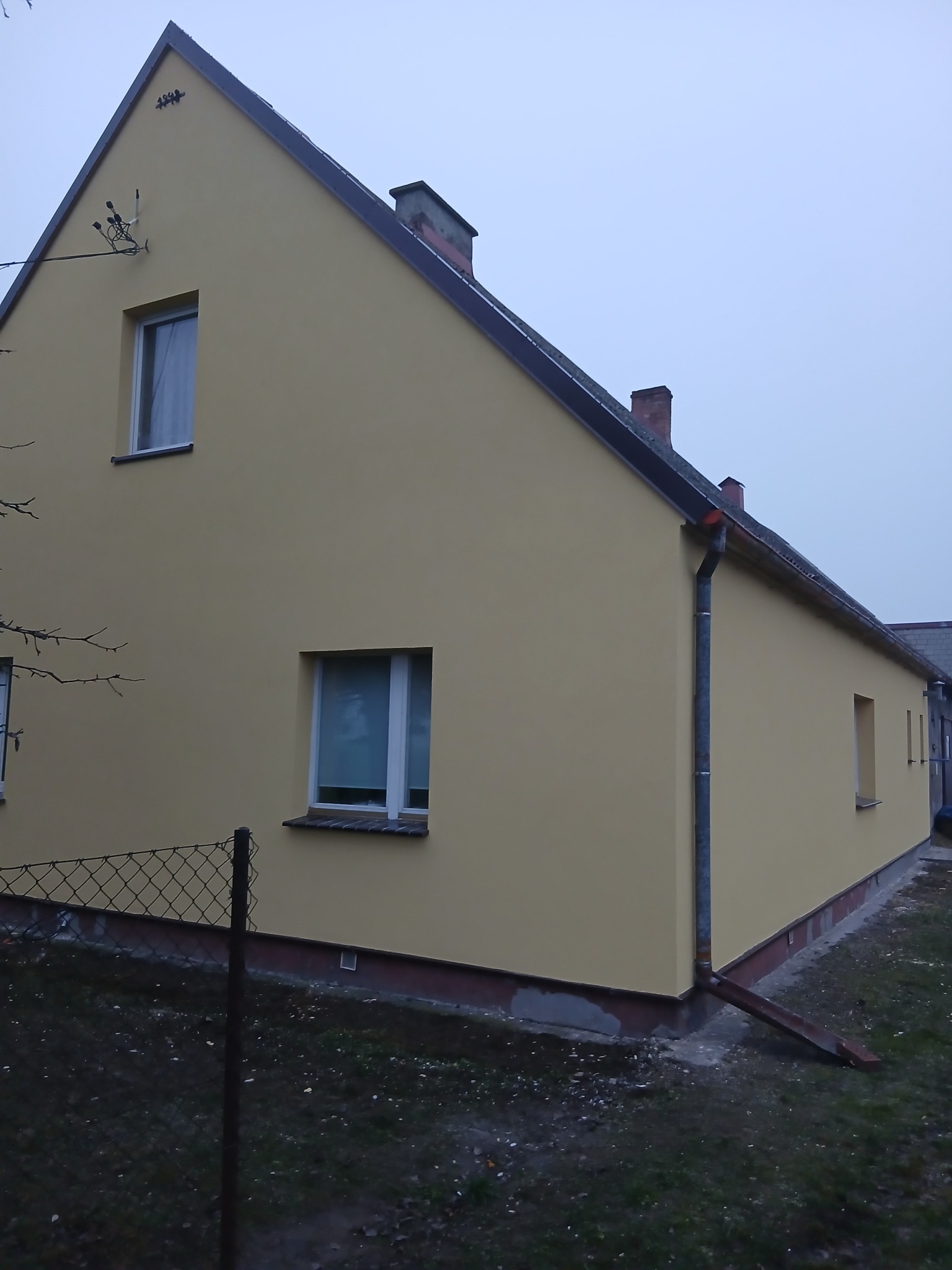 Żółta elewacja domu jednorodzinnego z białymi oknami i szarymi rynnami, widok z boku, ogrodzenie z siatki w tle, pochmurne niebo.