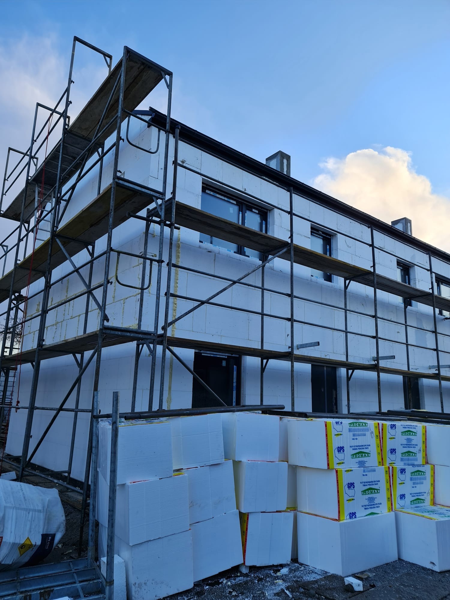 Budynek w trakcie ocieplania styropianem, widoczne rusztowania i paczki styropianu. Ściany budynku pokryte białymi płytami izolacyjnymi.