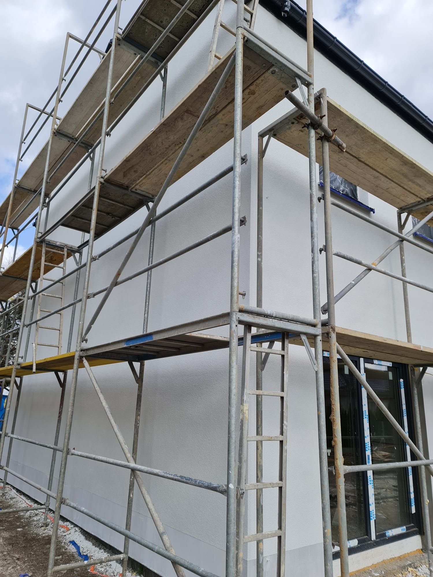 Elewacja domu w trakcie prac wykończeniowych, widoczne rusztowania i świeżo nałożony tynk strukturalny. Okna zabezpieczone folią. Detal: czarna rynna.