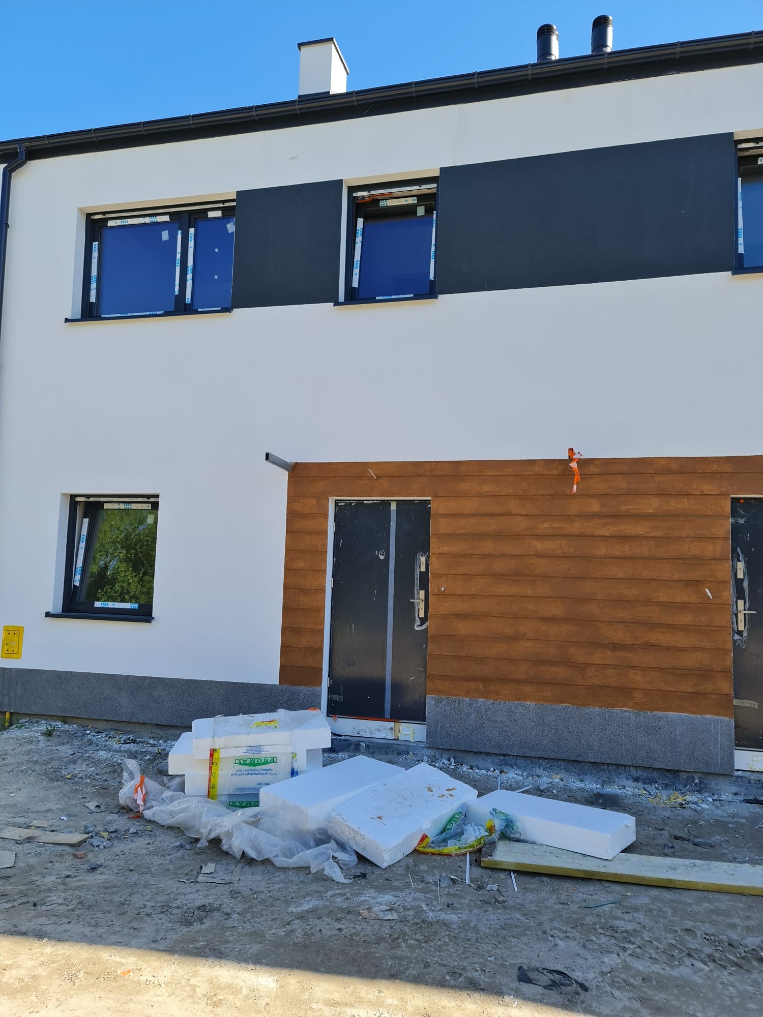 Nowoczesny dom jednorodzinny w trakcie ocieplania, z widocznymi płytami styropianowymi i fragmentami elewacji w kolorze białym, szarym i drewnopodobnym.