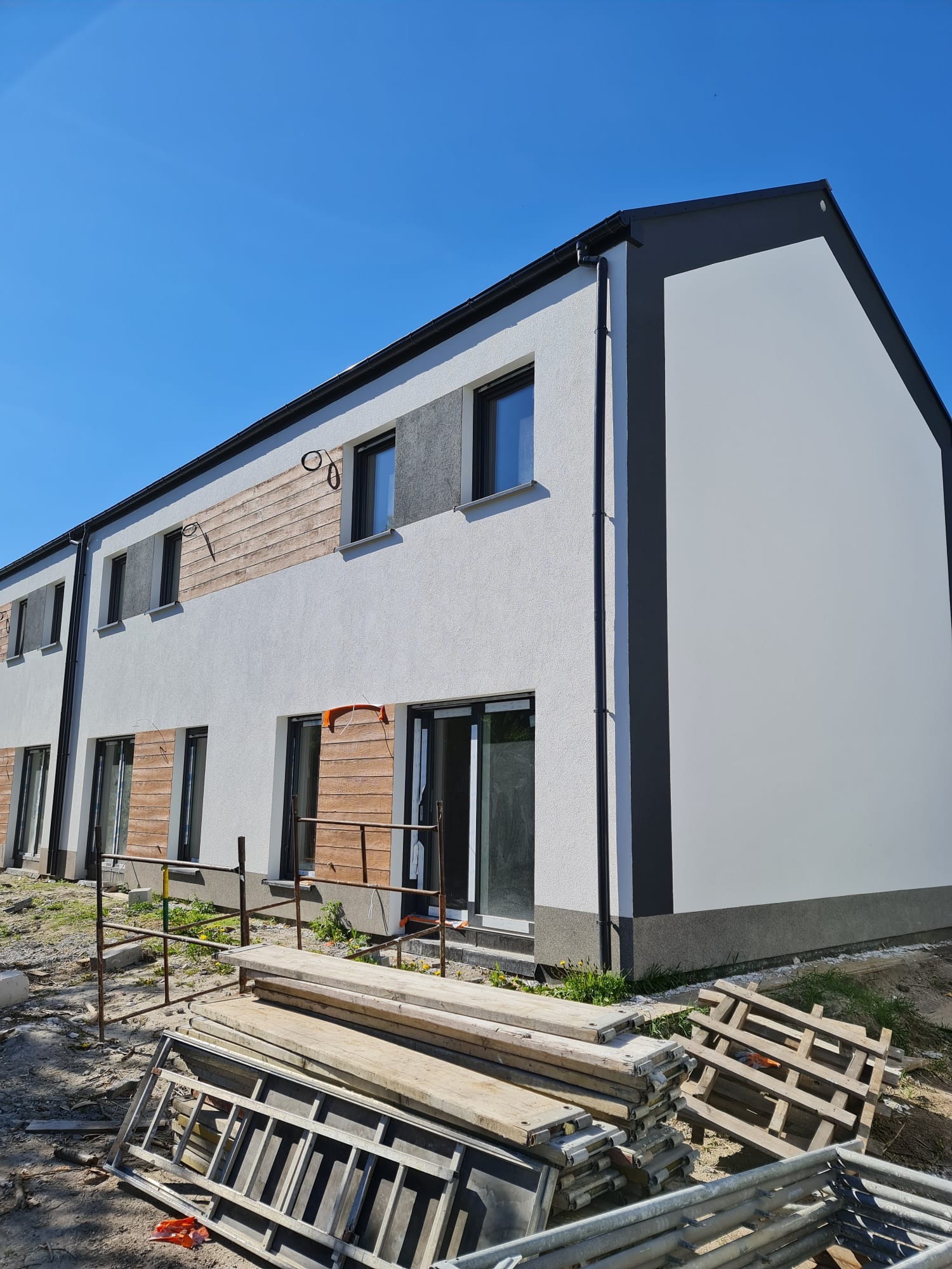 Nowoczesny budynek po ociepleniu, z fragmentami drewnianej elewacji i rusztowaniami w tle, w słoneczny dzień. Widoczne palety i drabina.