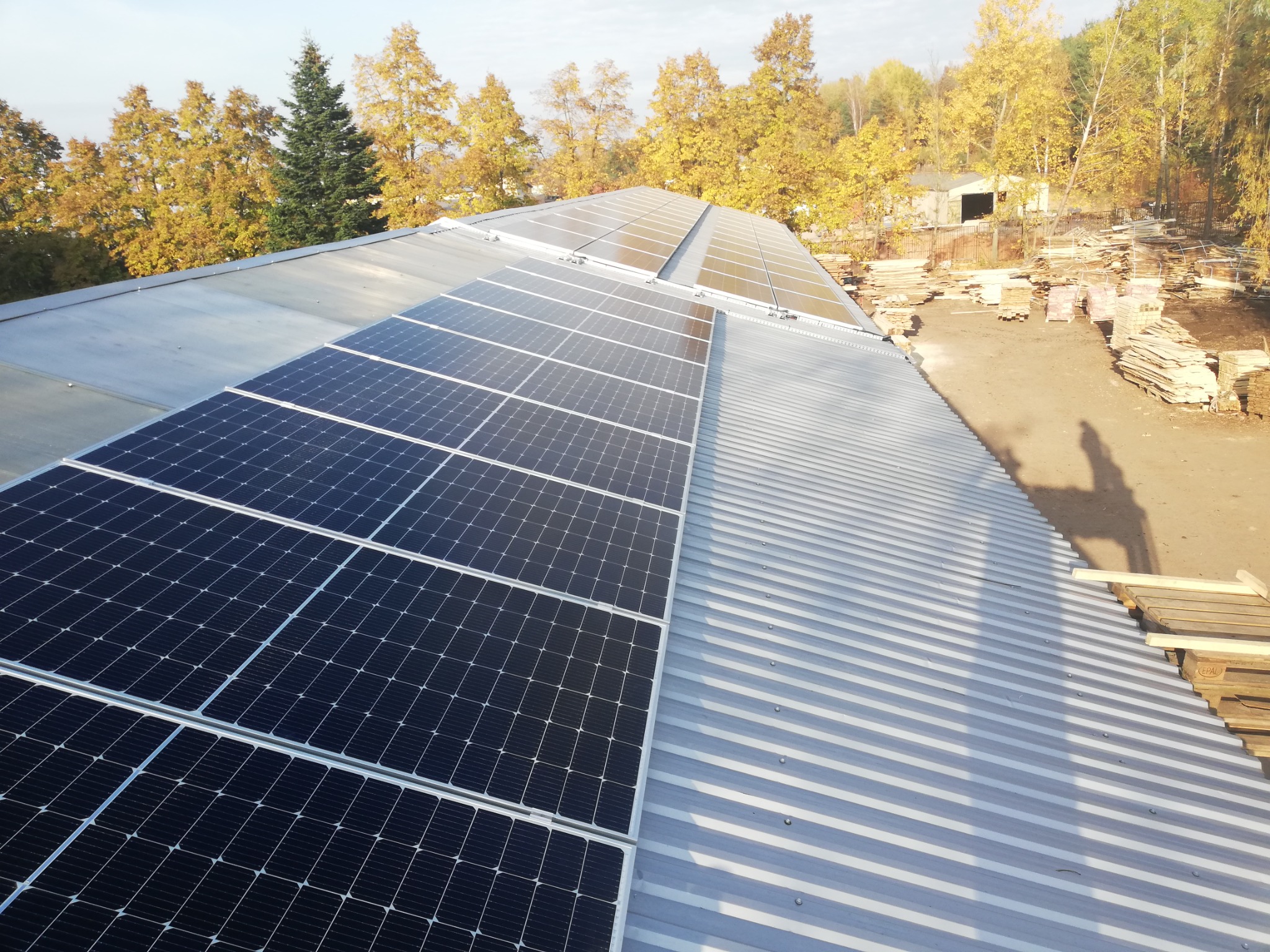 Panele fotowoltaiczne na blaszanym dachu tartaku, w tle składowane drewno i jesienne drzewa.