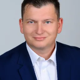 Portret uśmiechniętego mężczyzny w granatowej marynarce i białej koszuli, na jasnoszarym tle, z delikatnym cieniem pod brodą, sugerujący profesjonalizm i zaufanie.