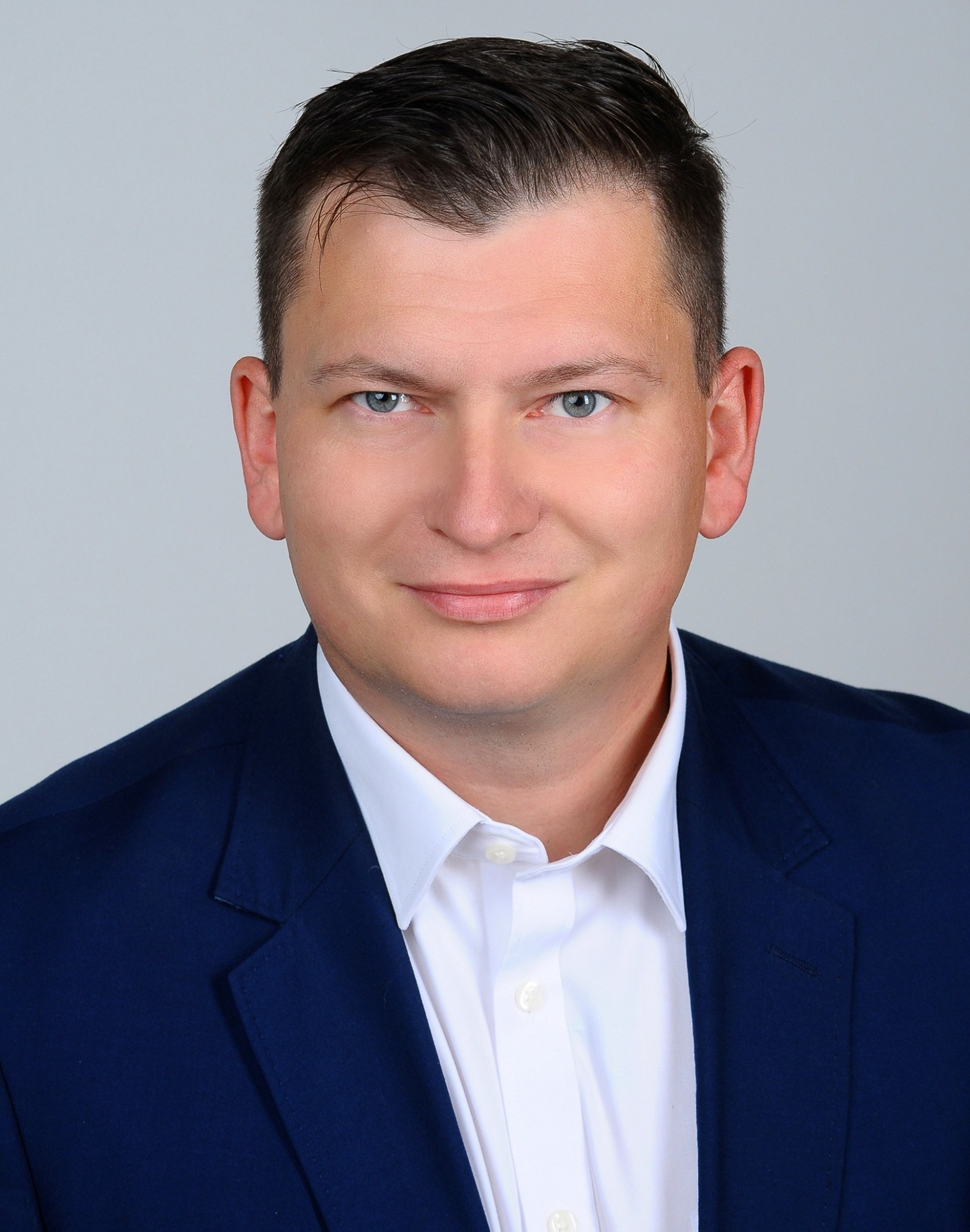 Portret uśmiechniętego mężczyzny w granatowej marynarce i białej koszuli, na jasnoszarym tle, z delikatnym cieniem pod brodą, sugerujący profesjonalizm i zaufanie.