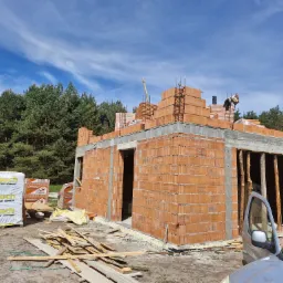 Wznoszenie ścian domu z czerwonej ceramiki, widoczny murarz na rusztowaniu, palety z kominami systemowymi Hoch i dachówką ceramiczną, teren budowy, las w tle i fragment samochodu.