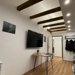 Pokój na poddaszu z drewnianymi belkami stropowymi, telewizorem na ścianie, stolikiem z cienkimi nogami i wieszakiem na ubrania w przejściu.