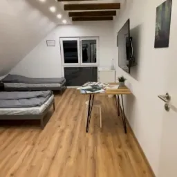 Pok&oacute;j na poddaszu z dwoma ł&oacute;żkami z szarą pościelą, drewnianą podłogą, biurkiem z laptopem i telewizorem na ścianie, oświetlony światłem z okna dachowego.