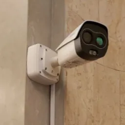 Kamera termowizyjna zamontowana na ścianie z dekoracyjną strukturą, widoczne przewody w listwie.