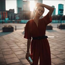 Radosław Kwasiborski - Kobieta w czerwonym kombinezonie z czarnym paskiem pozuje na tle miejskiej architektury w promieniach zachodzącego słońca, trzymając w dłoni małą książeczkę.