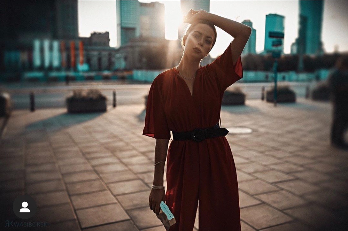 Kobieta w czerwonym kombinezonie z czarnym paskiem pozuje na tle miejskiej architektury w promieniach zachodzącego słońca, trzymając w dłoni małą książeczkę.