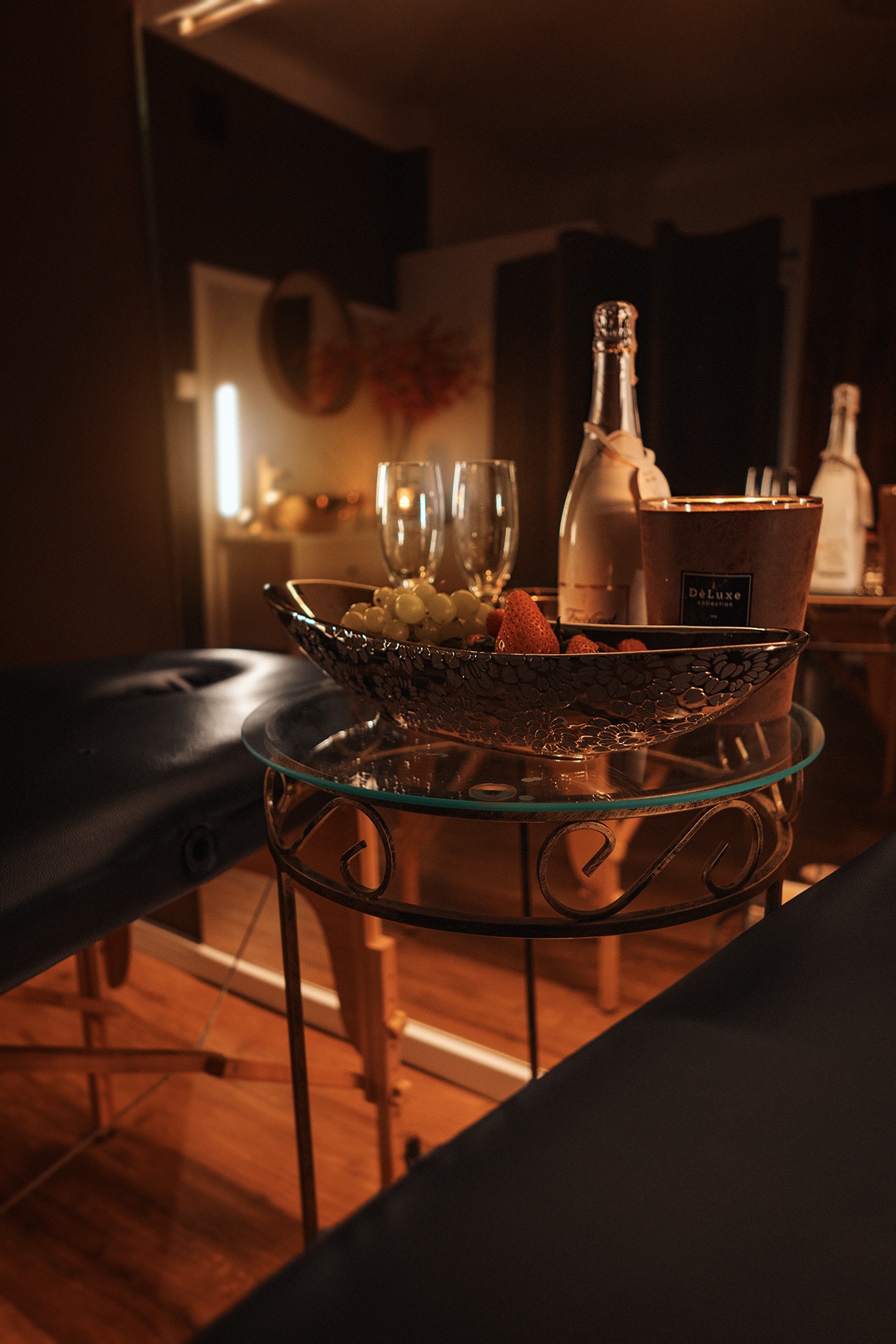 Przyciemnione wnętrze salonu z dwoma stołami do masażu, szampanem, owocami i świeczkami na szklanym stoliku z metalową podstawą.