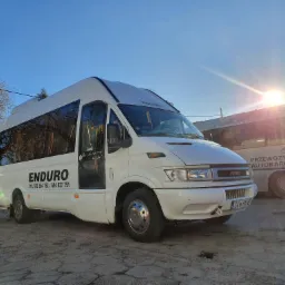 Biały bus marki Iveco z napisem 'ENDURO' i numerami telefonów na boku, stojący na placu w słoneczny dzień, obok zaparkowany autobus z napisem 'Przewozy Autokarowe', widoczne drzewa bez liści w tle.
