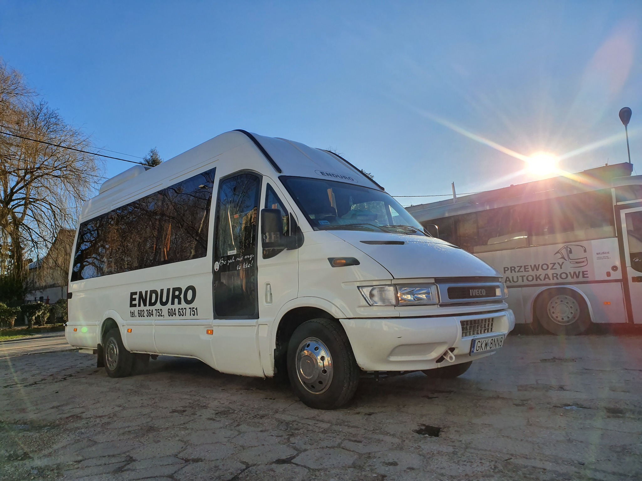 Biały bus marki Iveco z napisem 'ENDURO' i numerami telefonów na boku, stojący na placu w słoneczny dzień, obok zaparkowany autobus z napisem 'Przewozy Autokarowe', widoczne drzewa bez liści w tle.