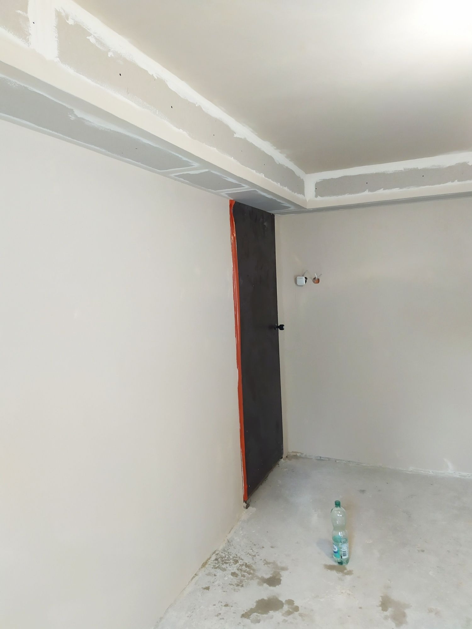 Wnętrze w trakcie prac wykończeniowych: ściany po szpachlowaniu, przygotowane do malowania, widoczne profile sufitowe i zabezpieczone drzwi, butelka wody na podłodze.
