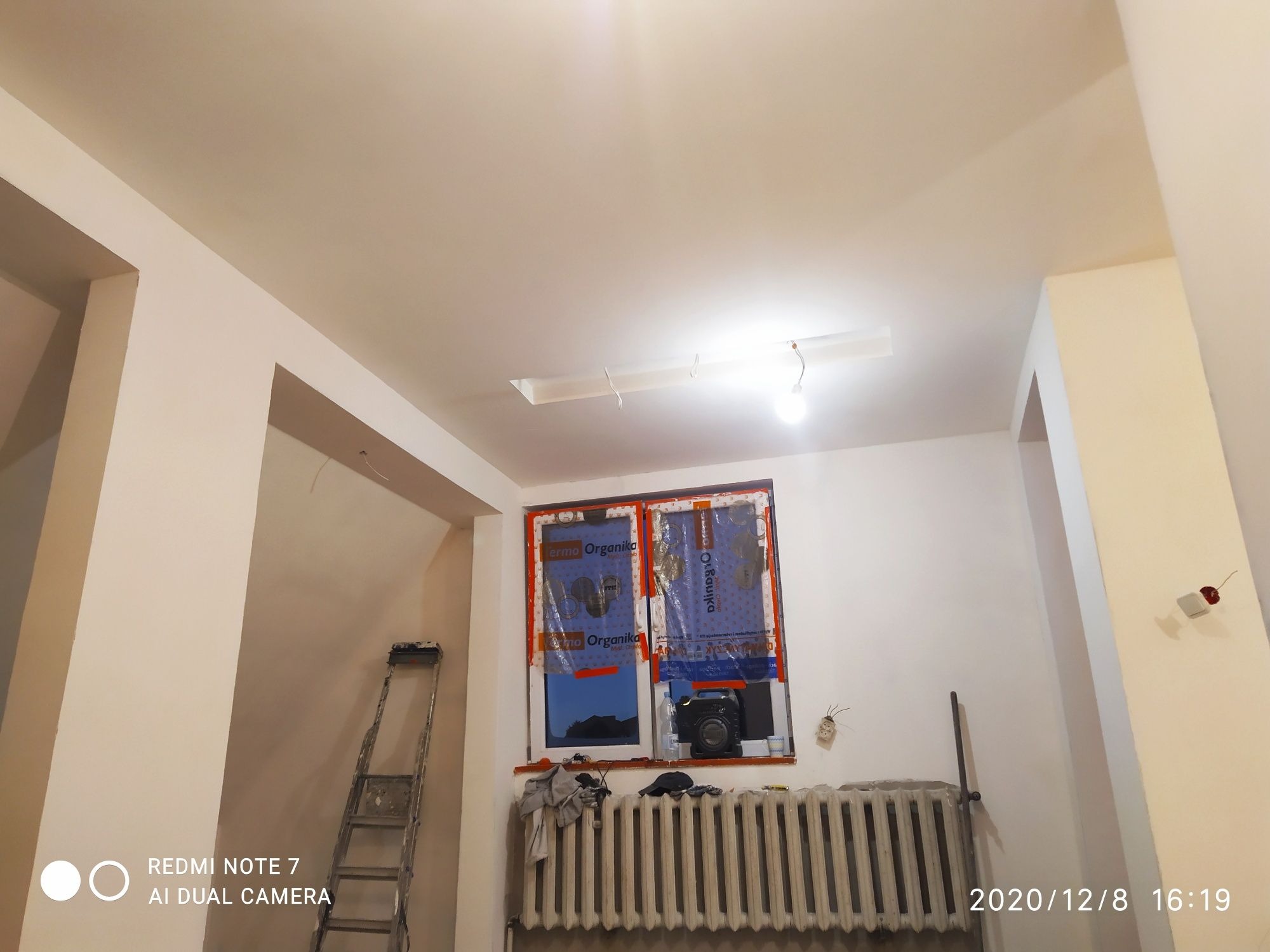 Wnętrze w trakcie prac wykończeniowych: świeżo pomalowane ściany w odcieniach bieli, zabezpieczone okno folią, drabina oparta o ścianę, grzejnik pod oknem