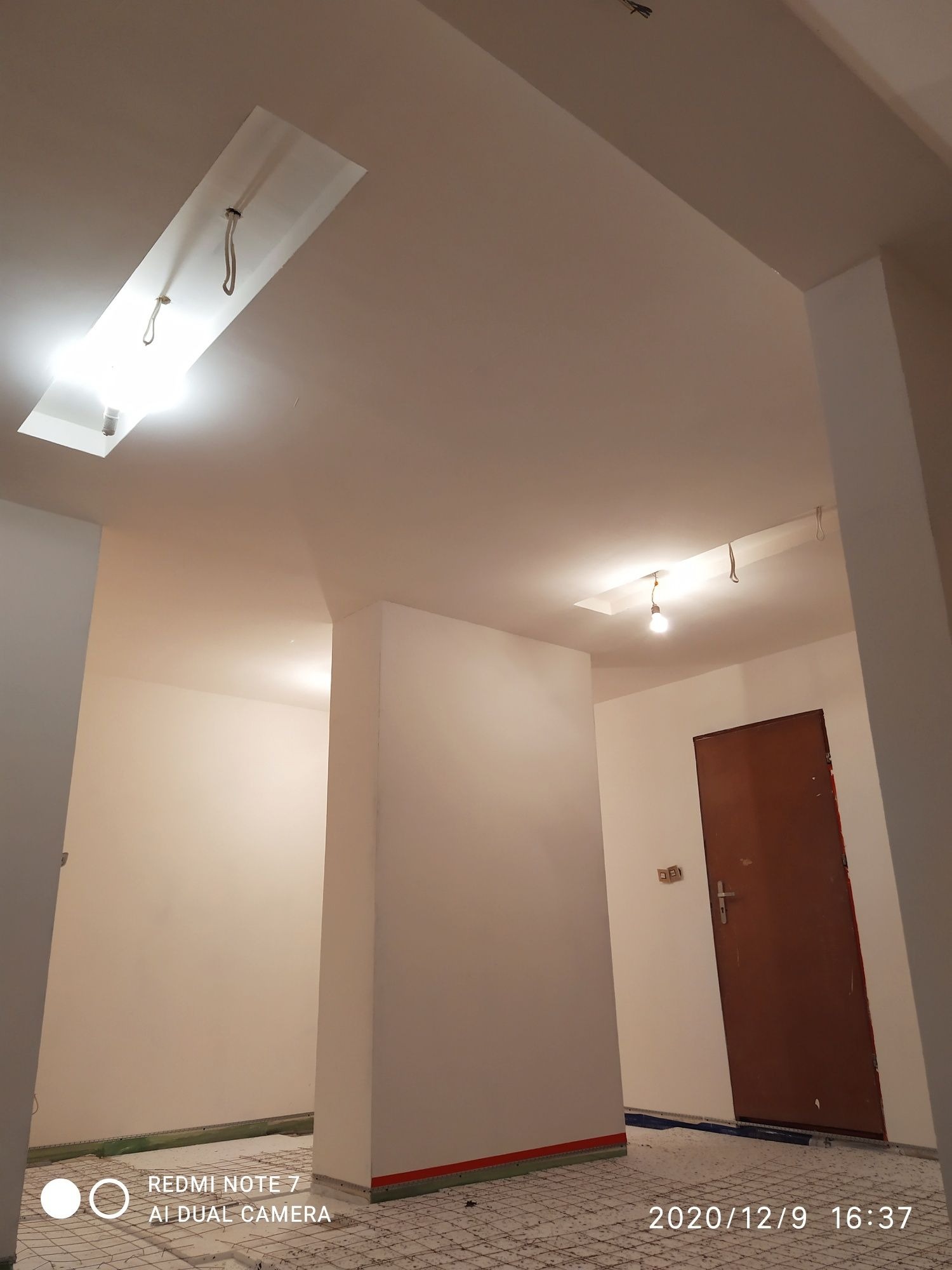 Wnętrze w trakcie prac wykończeniowych: świeżo pomalowane ściany na biało, widoczne instalacje oświetleniowe w suficie, podłoga zabezpieczona siatką i folią.