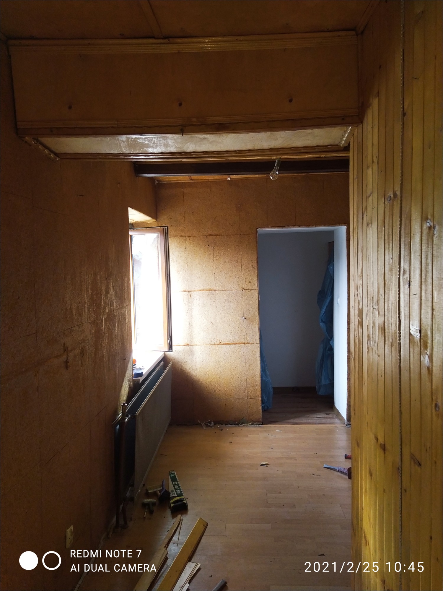 Wnętrze pokoju w trakcie prac remontowych, ściany wyłożone płytami, widoczne narzędzia i materiały budowlane, drewniana podłoga, okno z grzejnikiem, surowe wykończenie.