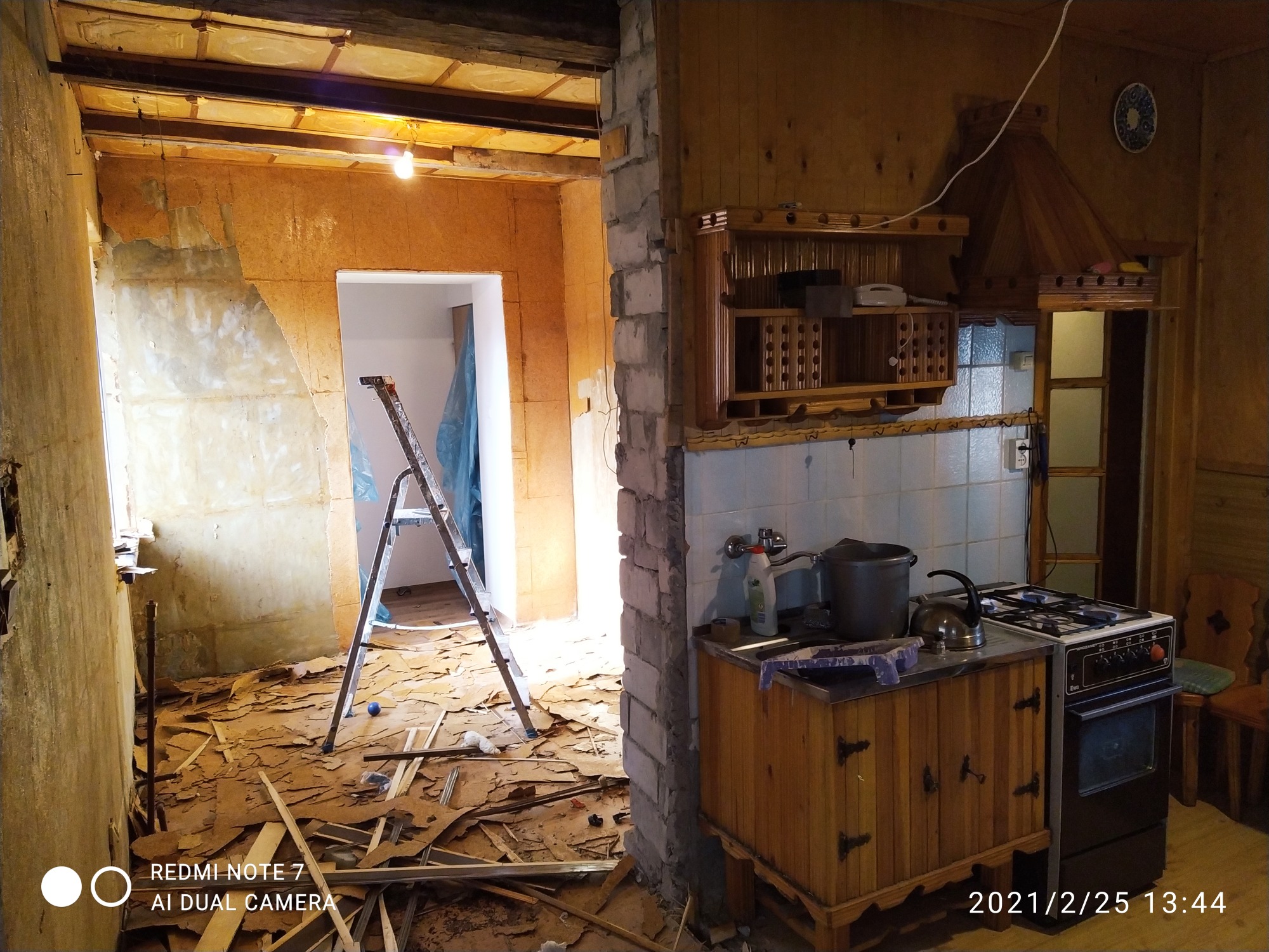 Kuchenne wnętrze w trakcie gruntownego remontu: zerwane stare panele podłogowe, odsłonięte fragmenty ścian, drabina rozstawiona w przejściu, widoczna stara zabudowa kuchenna z kuchenką gazową.