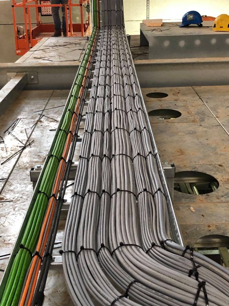 Rzędy kabli w korytach kablowych, uporządkowane za pomocą opasek zaciskowych, widoczne kable w kolorach zielonym, pomarańczowym i szarym, w tle podnośnik koszowy i kaski ochronne.