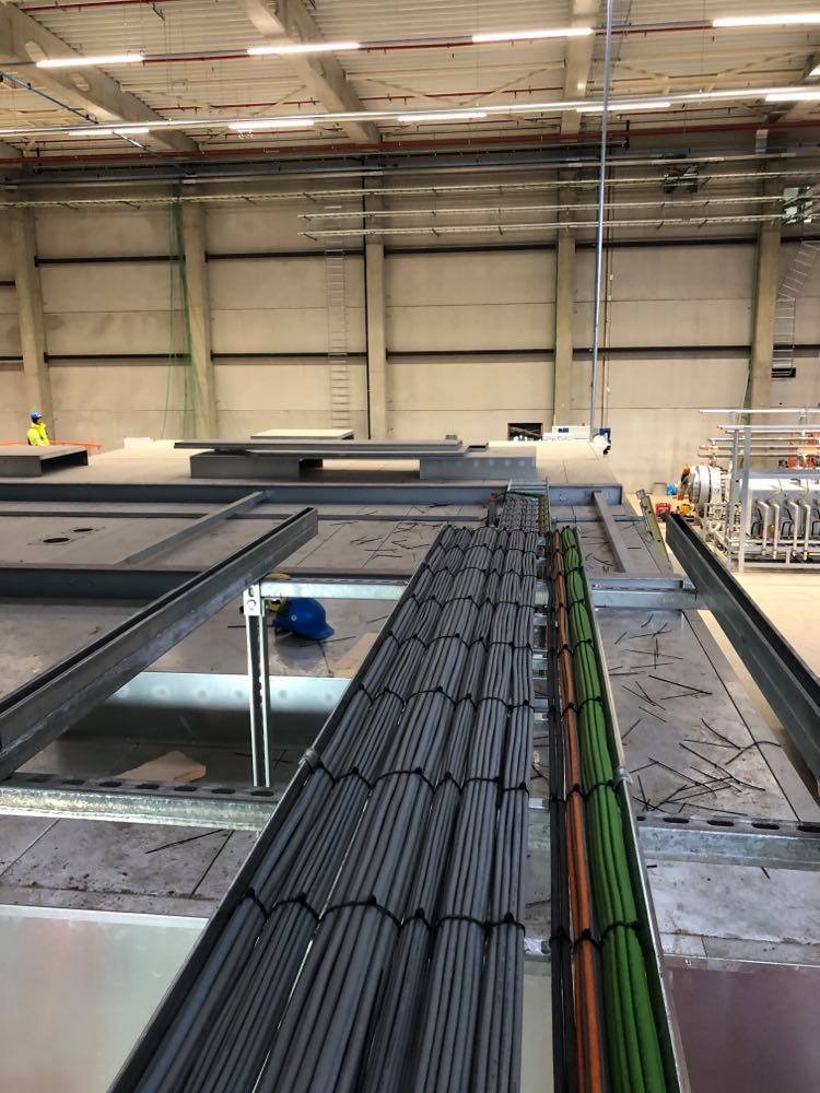 Kable elektryczne ułożone w korytach na konstrukcji wsporczej w hali przemysłowej, widoczny pracownik w oddali.