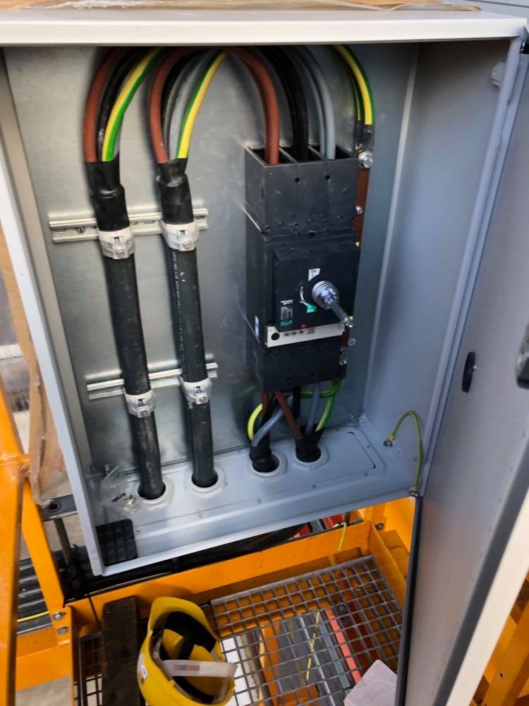 Otwarta skrzynka elektryczna z grubymi kablami w izolacji, wyłącznik prądu i uziemienie; żółty kask leży na metalowej kratce poniżej.