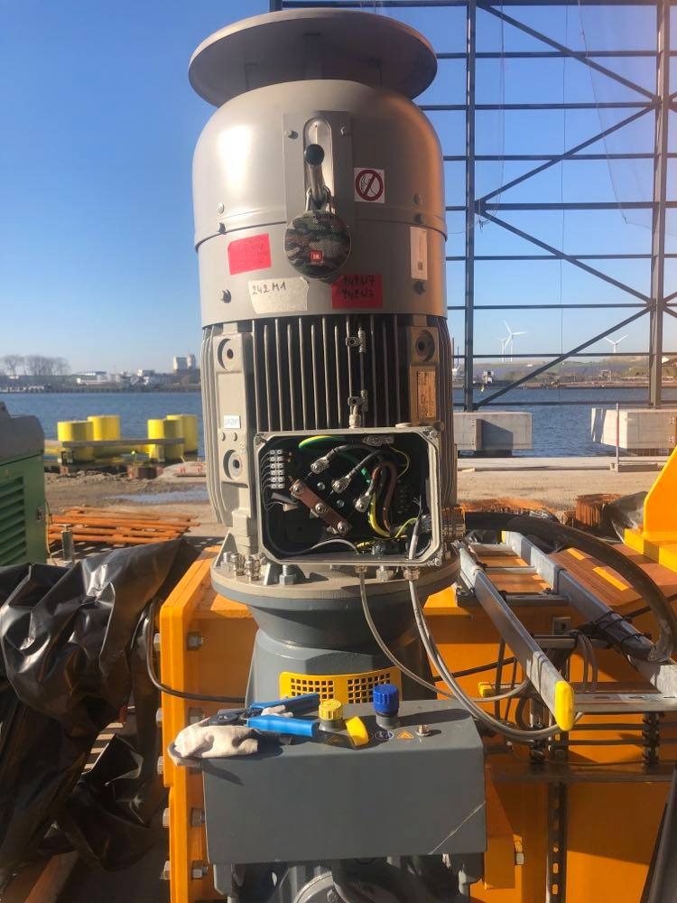 Otwarta skrzynka przyłączeniowa silnika elektrycznego z widocznymi przewodami, obok leżą narzędzia i rękawica robocza, w tle nabrzeże i wiatraki.