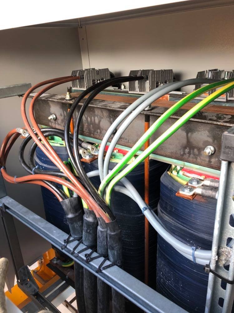 Wnętrze szafy elektrycznej z grubymi przewodami w różnych kolorach, podłączonymi do transformatorów, widoczne złącza i listwy zaciskowe.